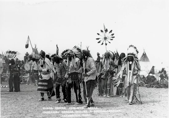 Indigenous people in traditional attire perform a dance outdoors, with tents in the background.