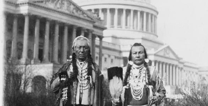 Two Native American men in traditional attire stand in front of a historical building.