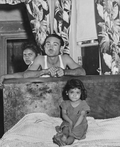 A man and a girl sit together on a bed, with a woman partially visible in the background.