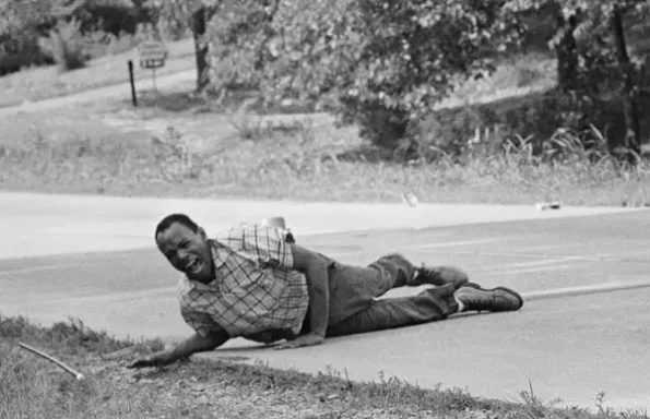 Man lying on the ground beside a road, appearing distressed or injured.